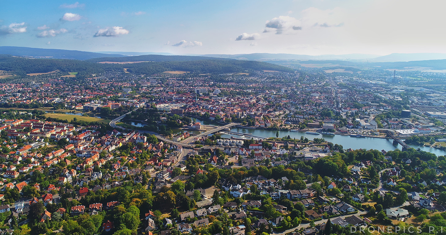 Hameln aus der Vogelperspektive: Die Stadt breitet sich mit ihren roten Dächern entlang der Weser aus. Mehrere Brücken überspannen den Fluss, der sich durch das Stadtgebiet schlängelt. Umgeben ist Hameln von grünen Hügeln, Wäldern und Feldern, die die Landschaft einrahmen.