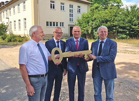 Vier Männer (v.l. Peter Hoffmann- Schönborn (BimA), Carsten Vetter (LK), Christian Mattern (GSW), Hermann Aden (Stadt Hameln)) stehen vor einem hellen Gebäude im Freien und halten gemeinsam einen großen symbolischen Schlüssel. Drei tragen Anzüge, einer ein kurzärmliges Hemd und Jeans. Hinter ihnen sind Bäume und ein teilweise gepflasterter Außenbereich zu sehen.